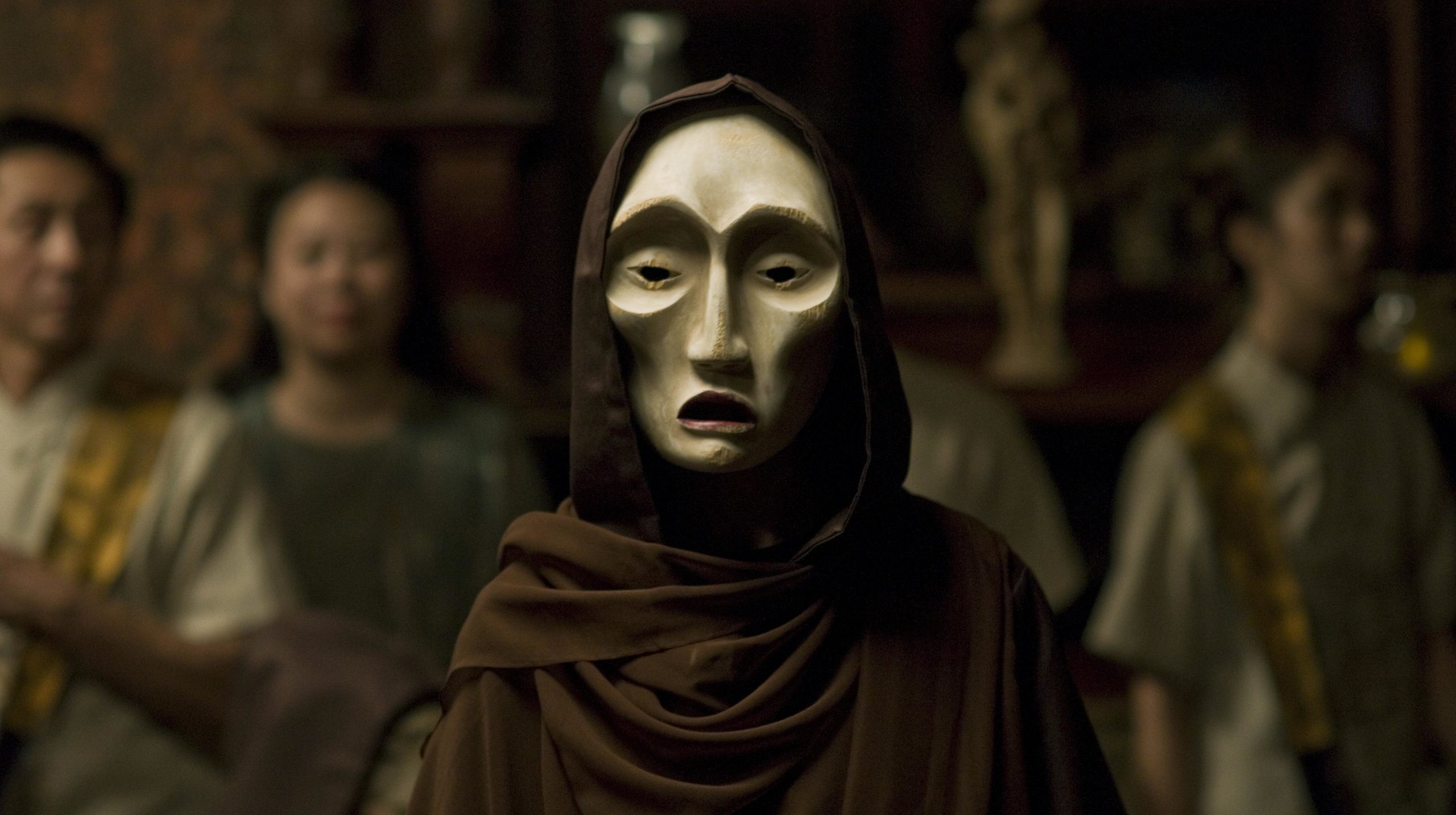 A still from the film <em>Drum Wave</em>: a figure wears a brown cloak and carved light wood mask