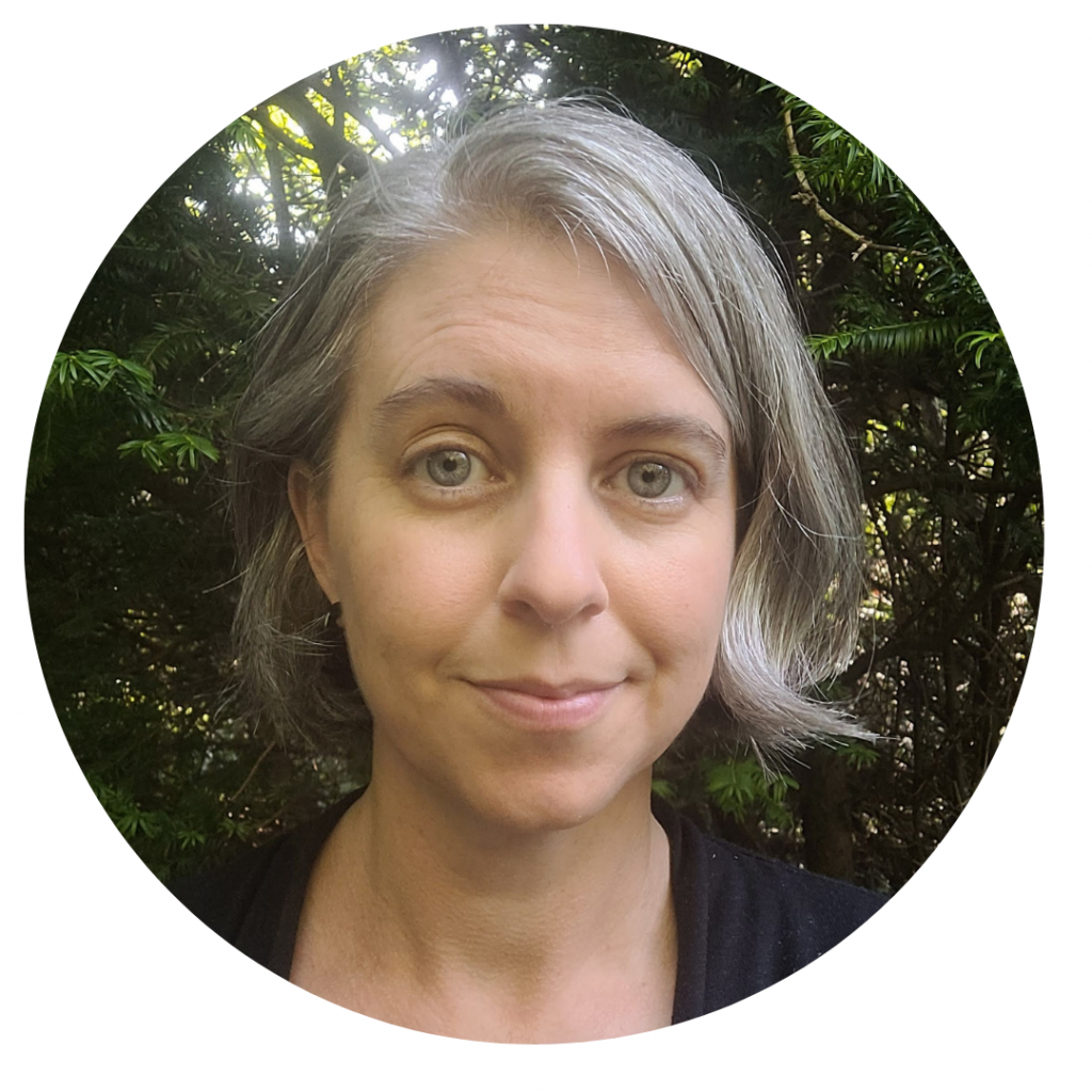 A photo of Erin Wiegand: a middle-aged white woman with short grey hair looks into the camera, smiling. She is wearing a black cardigan, and sunlight is coming through the trees behind her.